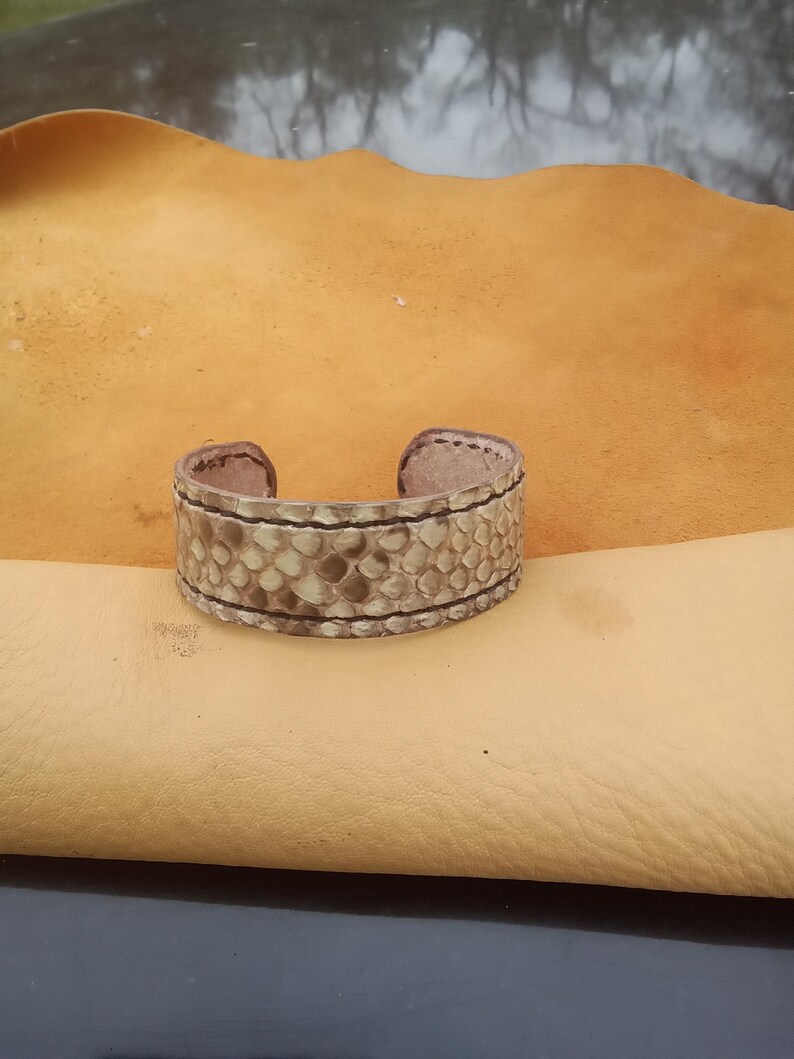 May include: A cuff bracelet made from textured, light-coloured leather with a reptile-like pattern. The bracelet has a rectangular shape with rounded edges and is accented with dark stitching. The bracelet is displayed on a tan leather surface.