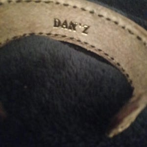 May include: A light tan leather bracelet with the name "DAN Z" embossed in silver lettering. The bracelet has a stitched border and is set against a dark blue background.