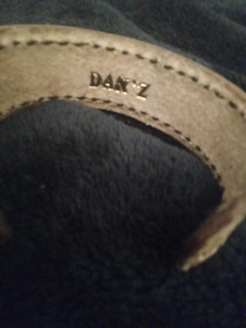 May include: A light tan leather bracelet with the name "DAN Z" embossed in silver lettering. The bracelet has a stitched border and is set against a dark blue background.