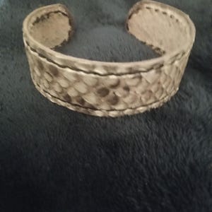 May include: A wide, open cuff bracelet with a snakeskin pattern in shades of grey and brown. The bracelet has a textured surface and is made of a flexible material. The edges are finished with darker stitching.