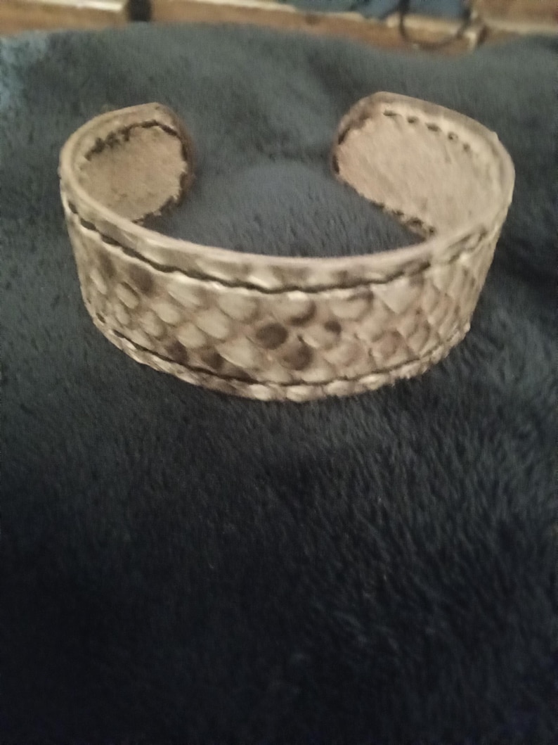 May include: A wide, open cuff bracelet with a snakeskin pattern in shades of grey and brown. The bracelet has a textured surface and is made of a flexible material. The edges are finished with darker stitching.