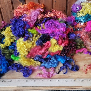 May include: A collection of colorful, curly wool roving in shades of blue, purple, pink, yellow, and green. Some roving is loose, while some is packaged in a clear bag. A ruler is placed on the wooden surface.