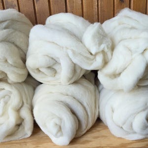 May include: Six individual bundles of off-white, unspun wool roving arranged in two rows of three. Each bundle is coiled into a soft, voluminous, and fluffy texture, showcasing the natural, fibrous appearance of the raw, unprocessed sheep wool material.