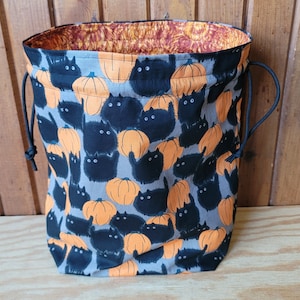 May include: A drawstring bag with a Halloween theme. The bag features a pattern of black cats and orange pumpkins on a gray background. The interior lining is a patterned orange and brown fabric. Black drawstrings are visible.