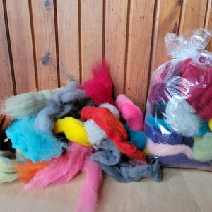 May include: A collection of colorful wool roving in various shades, including brown, teal, pink, and yellow. Some roving is loose, while a portion is contained in a clear plastic bag. The background features wooden planks.