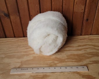 May include: A large, off-white ball of fluffy material, possibly batting or stuffing, sits on a light wood surface. A ruler with inch markings is placed below the ball, providing a size reference.