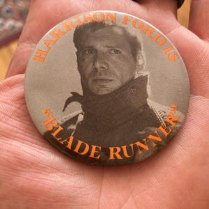 May include: A round button featuring a portrait of Harrison Ford from the movie "Blade Runner." The button has an orange border with the text "Harrison Ford is" and "Blade Runner."