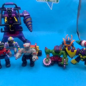 May include: A collection of colorful, vintage robot action figures. The robots are in various poses and designs, with a large purple robot in the background. The toys are on a blue surface.