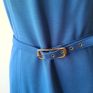 May include: A close-up of a blue dress with a matching belt. The belt has a gold-colored buckle and is secured with small gold-colored grommets. The dress has a textured appearance.