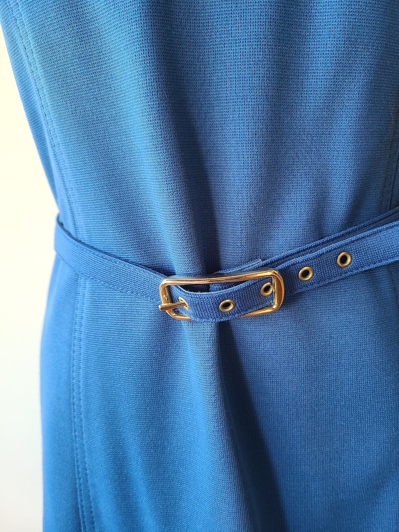 May include: A close-up of a blue dress with a matching belt. The belt has a gold-colored buckle and is secured with small gold-colored grommets. The dress has a textured appearance.