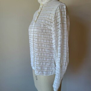 May include: White lace blouse with a high collar and long sleeves. The blouse has a ruffled collar and a button-up front.