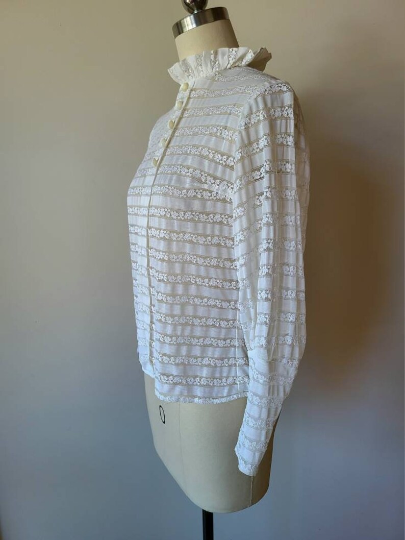 May include: White lace blouse with a high collar and long sleeves. The blouse has a ruffled collar and a button-up front.