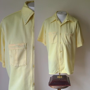 May include: A light yellow short-sleeved shirt with a pointed collar and two chest pockets. The collar and pockets are accented with green, red, and orange stitching. The shirt is button-down and displayed on a mannequin.