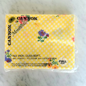 May include: A packaged flat top sheet by Cannon, featuring a yellow and white checkered pattern. The sheet is decorated with floral accents in shades of purple, orange, and green. The packaging includes text that reads "NO IRON DURA-SOFT" and "FULL FLAT".