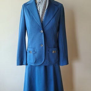 May include: A vintage teal skirt suit with a matching blazer and skirt. The blazer has gold-tone accents on the pockets and buttons. A striped scarf in white and navy blue is draped around the mannequin's neck. The skirt is a-line.
