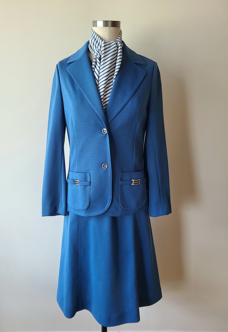 May include: A vintage teal skirt suit with a matching blazer and skirt. The blazer has gold-tone accents on the pockets and buttons. A striped scarf in white and navy blue is draped around the mannequin's neck. The skirt is a-line.