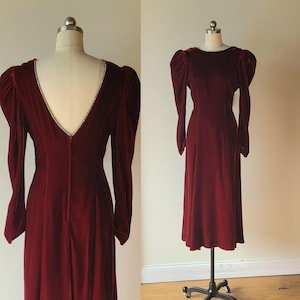 May include: A burgundy velvet dress with long sleeves and a floor-length skirt. The dress features a V-neckline trimmed with sparkling embellishments and dramatic puffed sleeves. The dress is displayed on a mannequin.