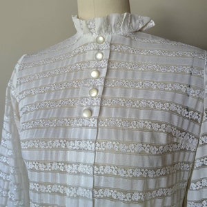 May include: White lace blouse with a high ruffled collar and a row of pearl buttons down the front.