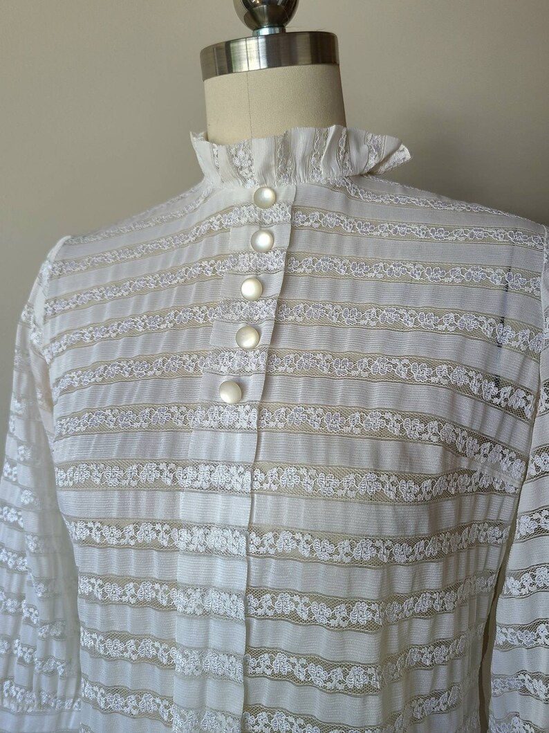 May include: White lace blouse with a high ruffled collar and a row of pearl buttons down the front.