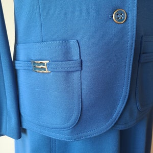 May include: A close-up of a bright blue suit jacket and skirt. The jacket features a gold button and a pocket with a gold-colored decorative accent. The fabric has a subtle texture. The skirt is visible at the bottom of the image.