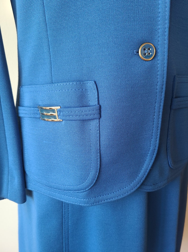 May include: A close-up of a bright blue suit jacket and skirt. The jacket features a gold button and a pocket with a gold-colored decorative accent. The fabric has a subtle texture. The skirt is visible at the bottom of the image.