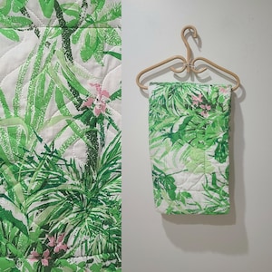 May include: A quilted textile with a vibrant tropical leaf pattern in shades of green and white, accented with small pink flowers. The textile is displayed on a natural wood hanger against a white background. The design features a repeating pattern of lush foliage.