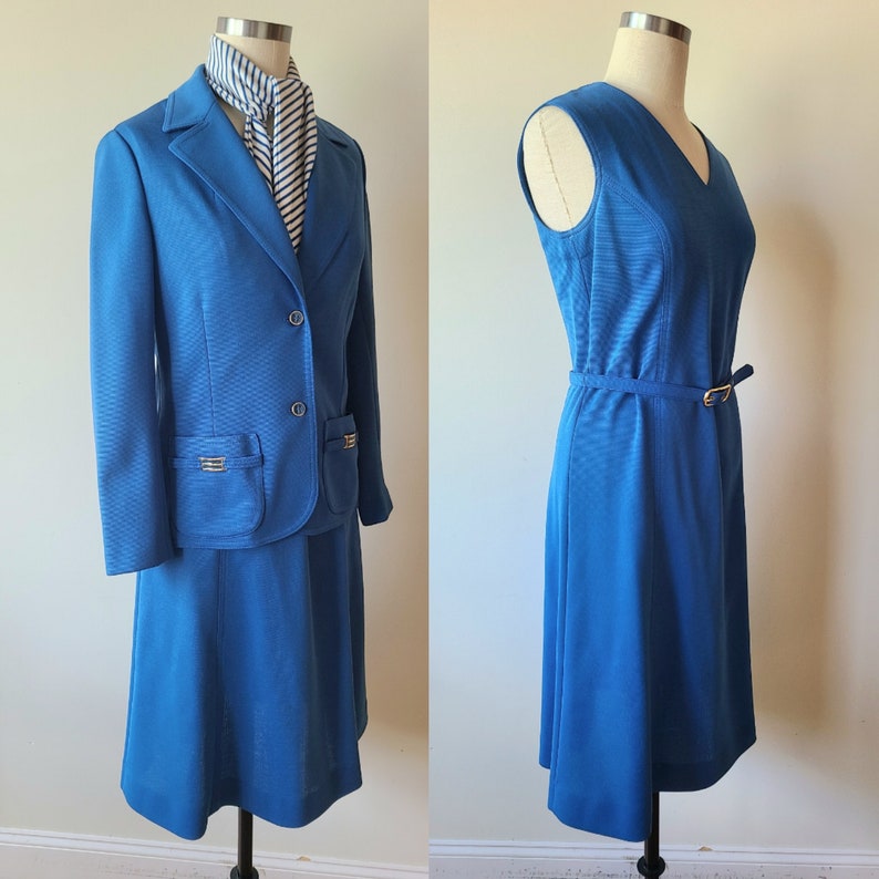 May include: Two-piece blue suit with a blazer, skirt, and a striped scarf. The blazer has two front pockets with gold-colored accents and matching buttons. A sleeveless blue dress with a belt and gold-colored buckle is also displayed.