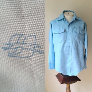 Puede incluir: Camisa abotonada azul claro con dos bolsillos en el pecho y cuello. La camisa tiene un patrón sutil de pequeños diseños azul claro. El lado izquierdo de la imagen muestra un primer plano de la tela con un diseño bordado en azul.