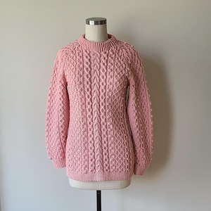 May include: A light pink sweater with a mock neck and intricate cable knit design. The sweater is displayed on a mannequin and features textured details on the sleeves and body. The overall style is classic and cozy.