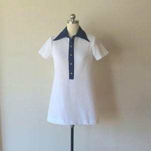 May include: White mini dress with a navy blue collar and placket. The dress has short sleeves and a button-down front. The dress is displayed on a mannequin. The dress is a vintage style.