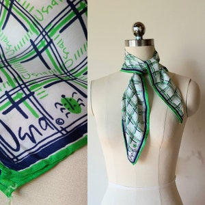 Peut inclure: Un foulard carré à fond blanc orné d'un motif à carreaux composé de lignes bleu foncé et vert vif. Le design comprend le texte "Usna" en écriture cursive et de petites illustrations de scarabées verts. Le foulard est noué autour d'un mannequin.