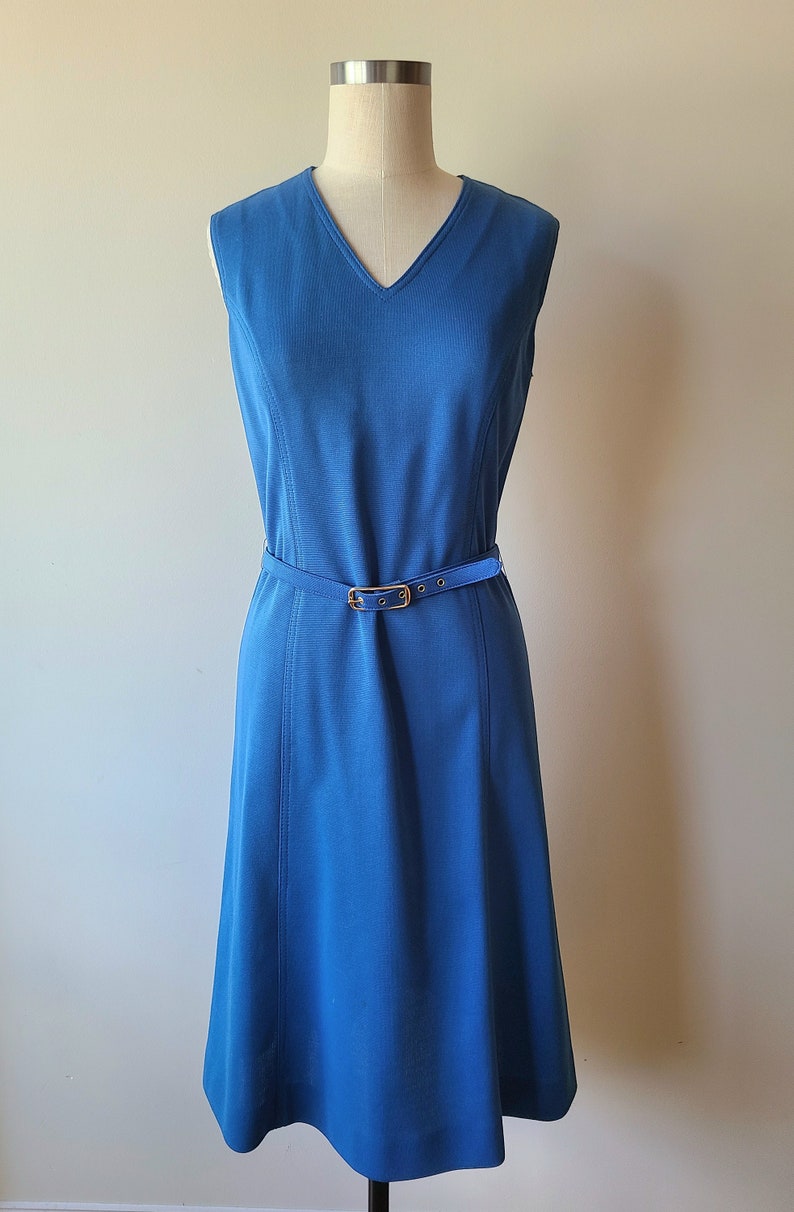 May include: A sleeveless, knee-length blue dress with a V-neck and a matching belt. The dress features vertical seams and a gold-colored buckle on the belt. The dress is displayed on a mannequin.