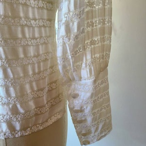 May include: White long-sleeved blouse with a striped pattern and lace detailing. The blouse has a buttoned cuff on the sleeve.