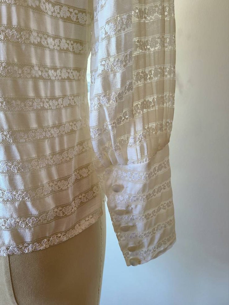 May include: White long-sleeved blouse with a striped pattern and lace detailing. The blouse has a buttoned cuff on the sleeve.