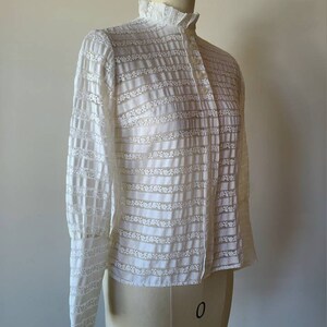May include: A white, long-sleeved blouse with a high collar and a button-up front. The blouse is made of a sheer, lace-like fabric with a striped pattern.