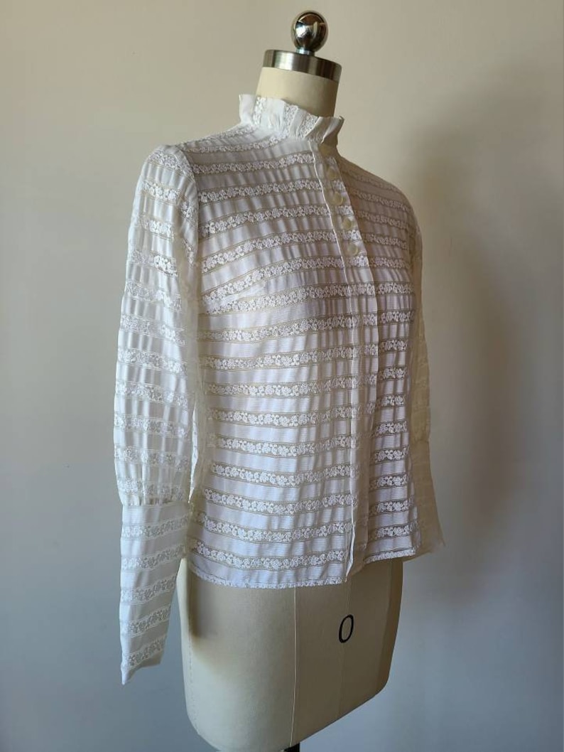 May include: A white, long-sleeved blouse with a high collar and a button-up front. The blouse is made of a sheer, lace-like fabric with a striped pattern.