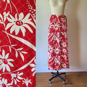 May include: A long, red skirt with a white floral design. The skirt has large white daisy-like flowers and long, white leaf shapes against a red backdrop. The skirt is displayed on a mannequin.