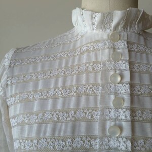 May include: White lace blouse with a high ruffled collar and a row of white buttons down the front.