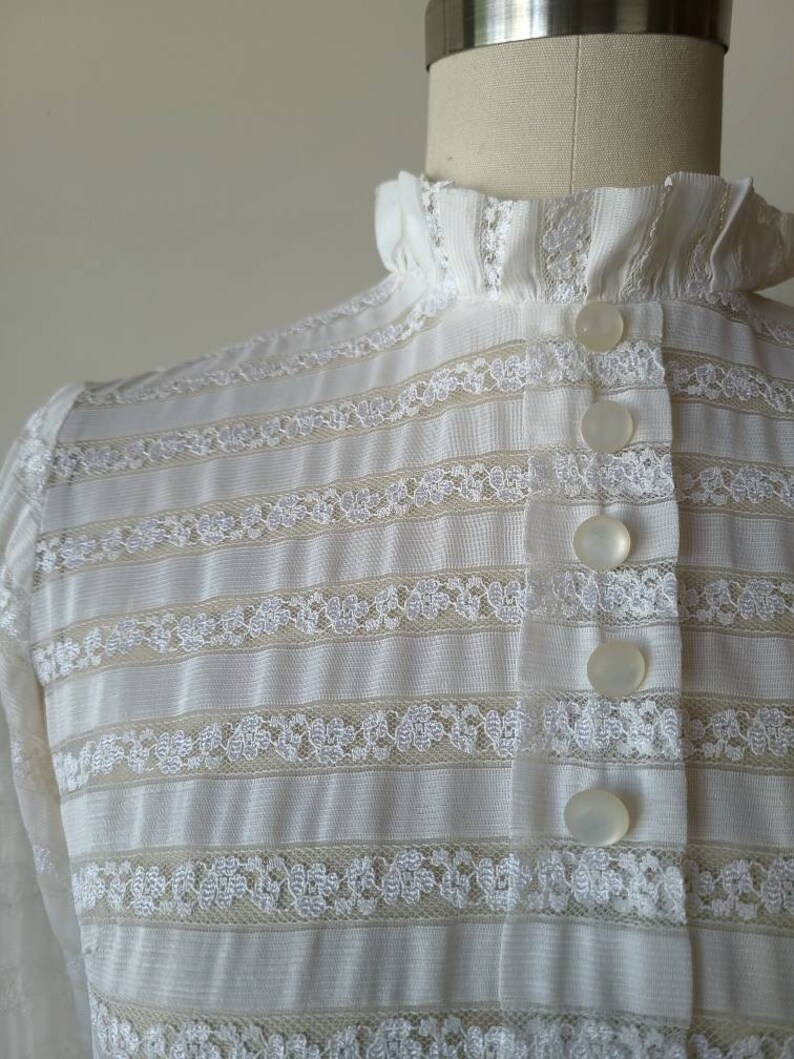May include: White lace blouse with a high ruffled collar and a row of white buttons down the front.