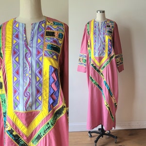 May include: A long, pink dress with a V-neck and long sleeves. The dress features colorful geometric patterns in yellow, green, purple, and blue. Gold trim accents the design. The dress is displayed on a mannequin.