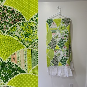 May include: A green and white patchwork quilt with a floral pattern and white lace trim. The quilt is hanging on a white hanger against a white wall.