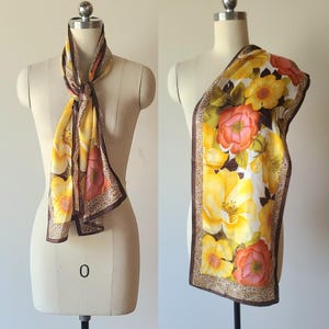 May include: A rectangular scarf featuring a vibrant floral pattern with large yellow and orange flowers on a white background. The scarf is bordered by a thin brown frame with a small leopard print detail and includes the text Oscar de la Renta.