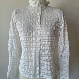 May include: White lace blouse with a high ruffled collar and long sleeves. The blouse has a button-up front and a striped pattern.