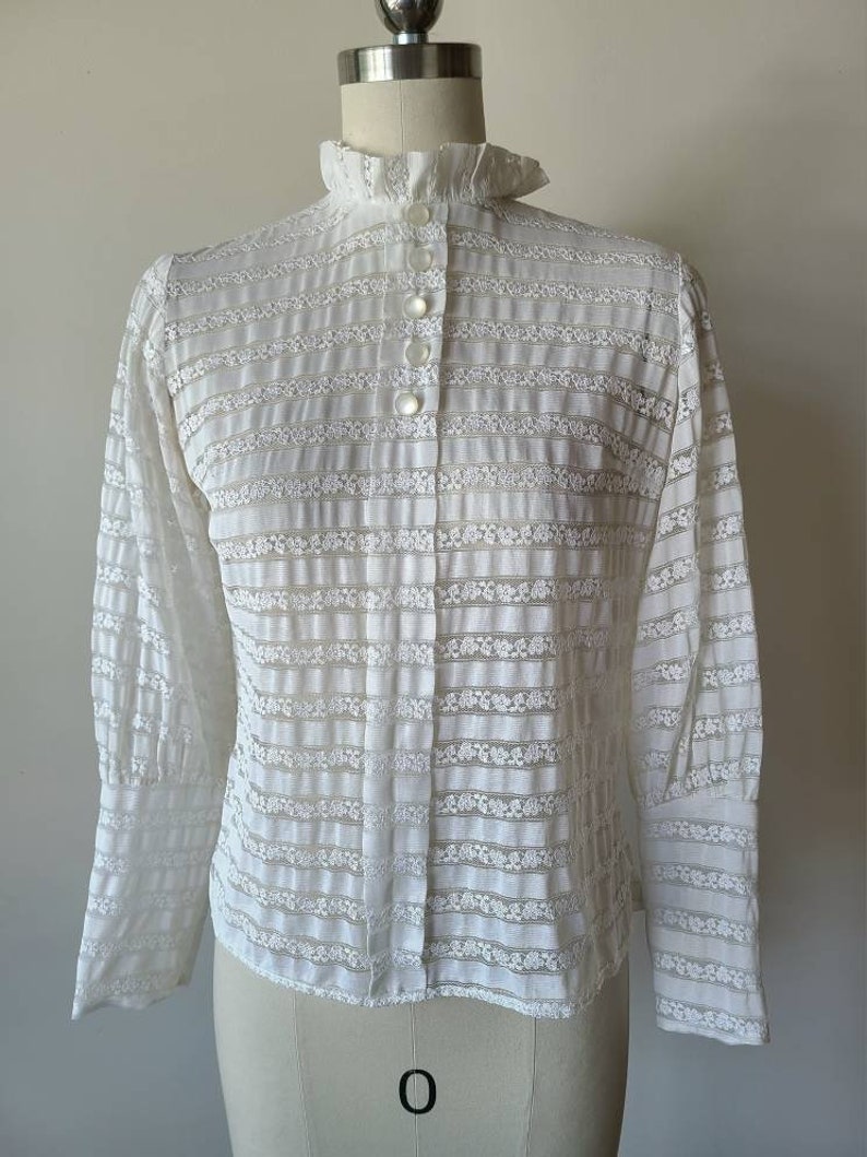 May include: White lace blouse with a high ruffled collar and long sleeves. The blouse has a button-up front and a striped pattern.