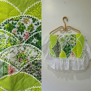 以下が含まれることがあります： 緑と白のパッチワーク生地に花柄が施されています。生地は白いレースで縁取られ、波打つような縁取りがされています。