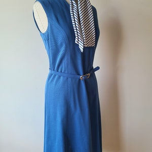 May include: A sleeveless, knee-length blue dress with a matching belt and gold buckle. A striped scarf in white and navy blue is draped over the mannequin. The dress is displayed on a black stand.