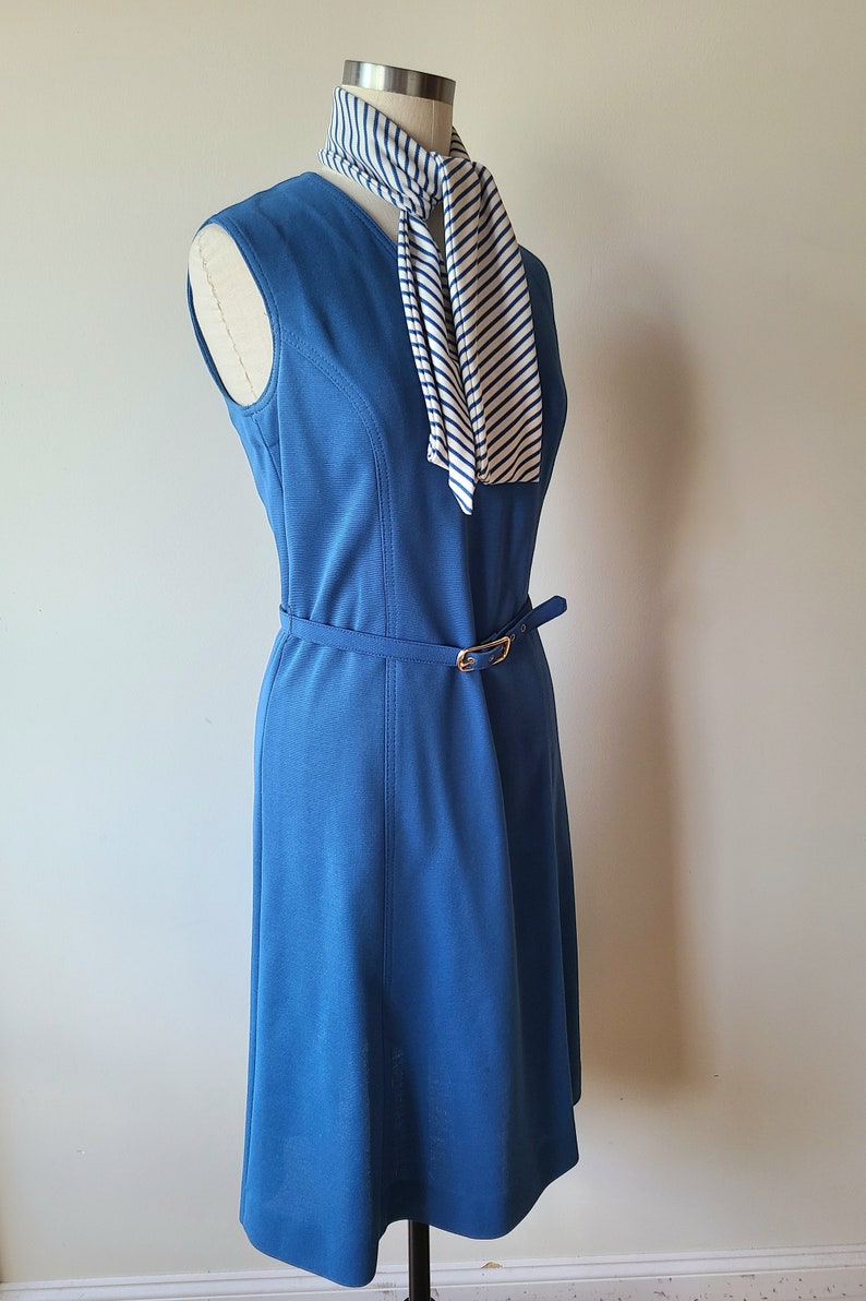 May include: A sleeveless, knee-length blue dress with a matching belt and gold buckle. A striped scarf in white and navy blue is draped over the mannequin. The dress is displayed on a black stand.