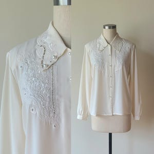 以下が含まれることがあります： 襟と胸元に複雑な白い花柄の刺繍が施された、オフホワイトの長袖ボタンダウンシャツ。尖った襟、カフス付きの長袖、そして小さな真珠のようなボタンで飾られたフロントのボタン留めが特徴の衣類です。
