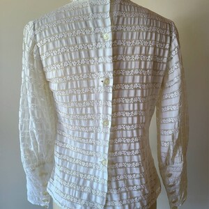May include: White long-sleeved blouse with a ruffled collar and a button-up front. The blouse is made of a sheer, delicate fabric with a floral pattern.