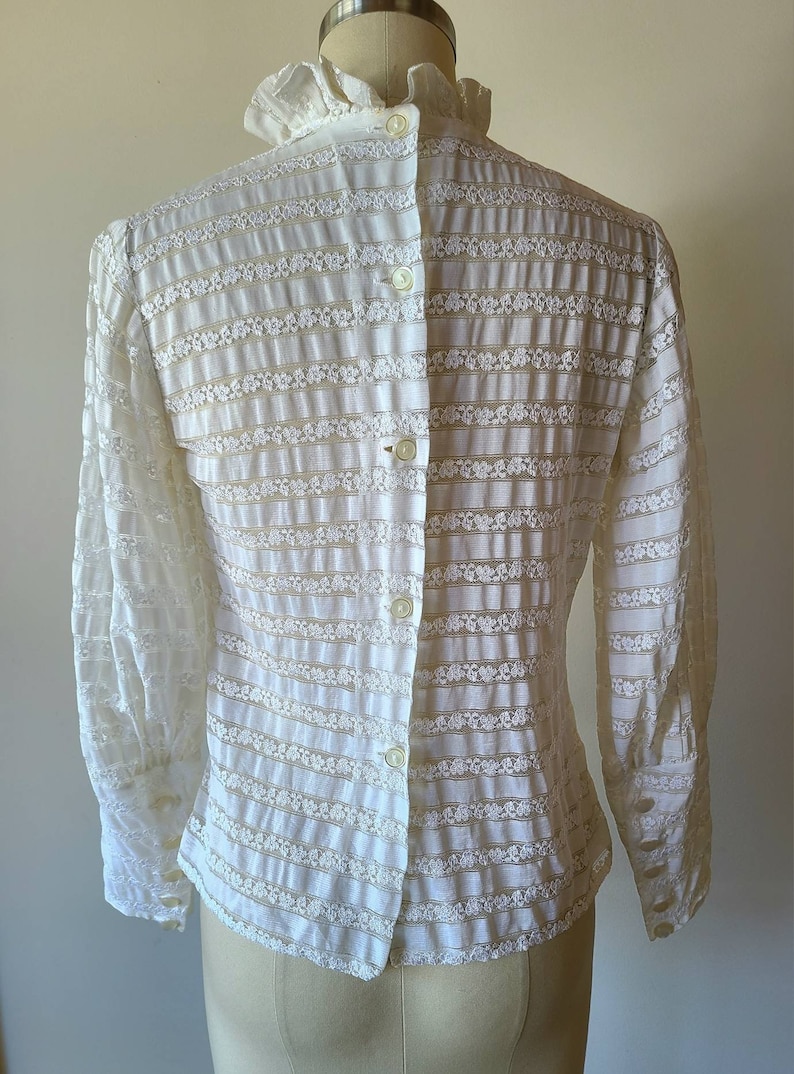 May include: White long-sleeved blouse with a ruffled collar and a button-up front. The blouse is made of a sheer, delicate fabric with a floral pattern.